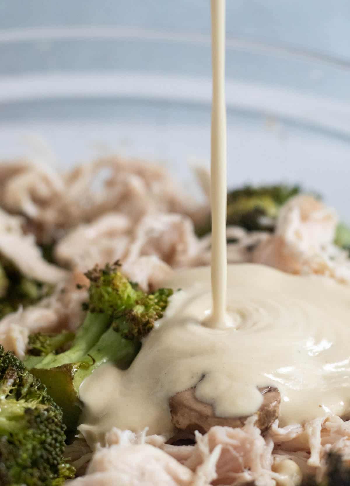 pouring alfredo sauce over chicken and broccoli in a clear bowl